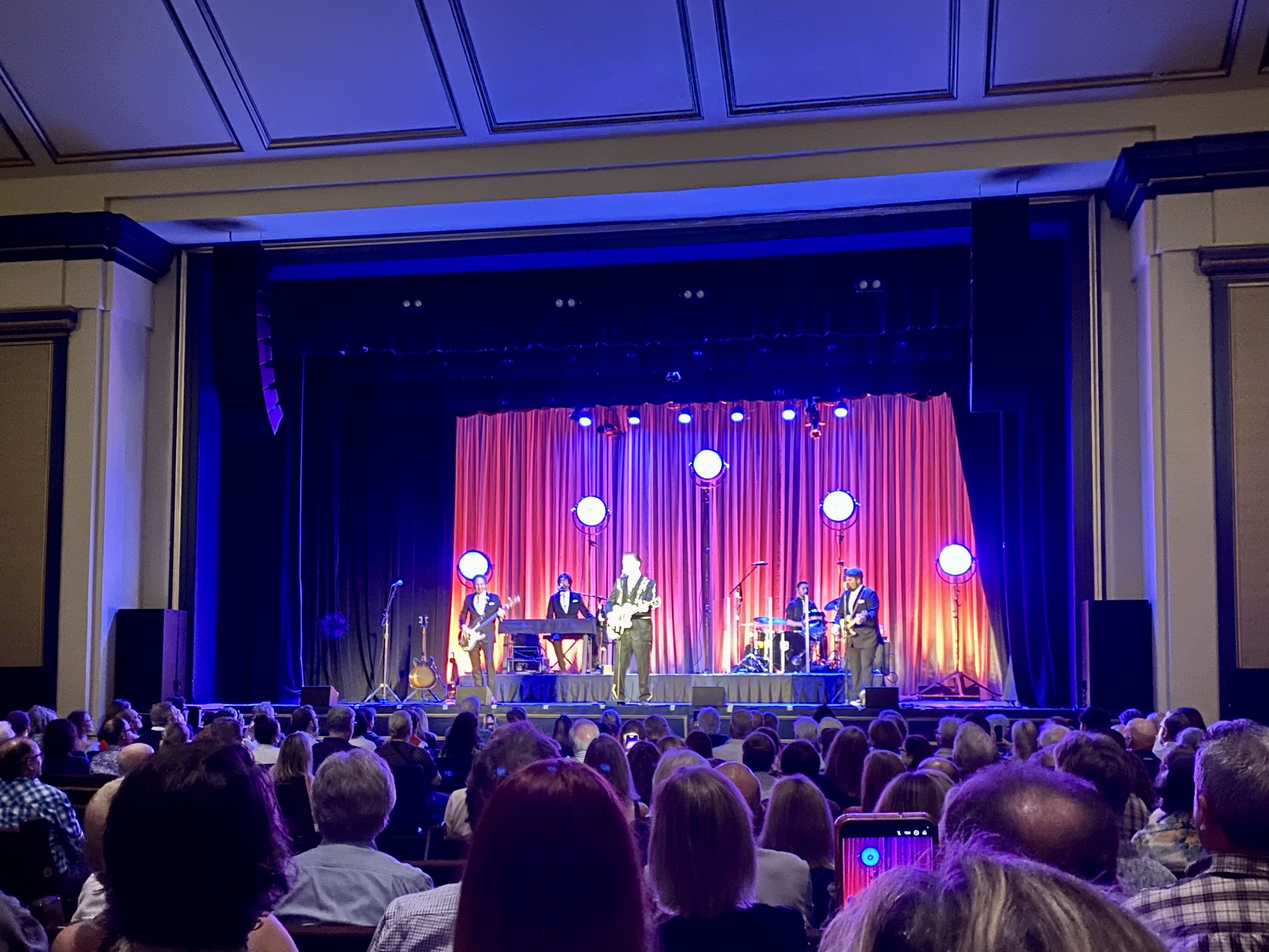 Chris Isaak on stage with his band, with onlooking audience. 