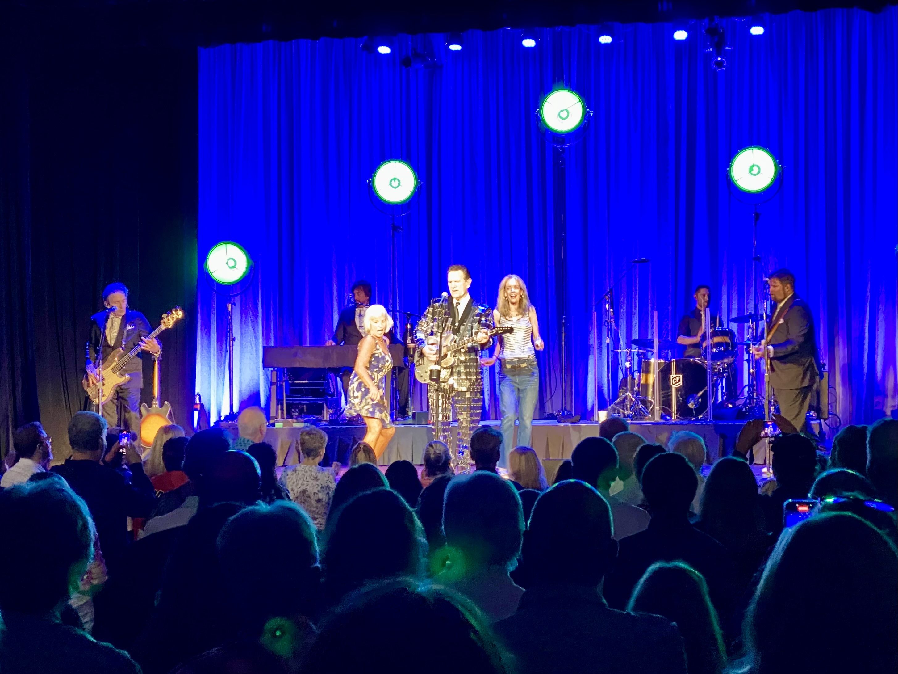 Chris Isaak sings on stage with two ladies from the audience dancing beside him. 